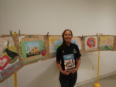 Dr. Micaela Rubalcava poses for a photo in the TMCC Main Gallery, proudly holding her new book and standing before her student-created recycled paper bag collage.