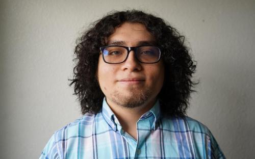 Jordi Cruz Trujillo stands for a photograph with a plain white backdrop wearing an aquamarine, baby, and denim blue plaid shirt.