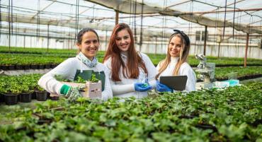 Three Agricultural Science students stand smiling in a lush greenhouse with hundreds of potted plants, a biological microscope, a tablet computer, test tubes, and a beaker filled with emerald-hued fluid.