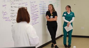 Two nursing students, one in black scrubs and the other in green, smile and laugh while learning detailed steps to coordinate provider's orders and home medications listed on a whiteboard across from their instructor.