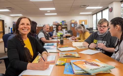 Dr. Micaela Rubalcava smiles at the camera, sitting at a lengthy beige desk with her students alongside children's books, colored pencils, and paper.