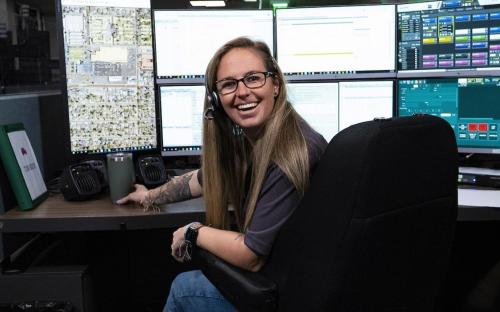 An emergency dispatch responder smiles while sitting behind her six computer screens, monitoring incoming or outgoing calls.