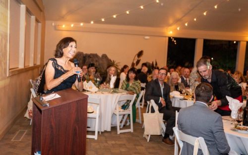 Dr. Cecilia Vigil gives her acceptance speech to a crowd sitting in white chairs and tables with strings of warmly lit bulbs above.