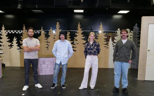 Four theatre students pose, smiling on a "Nearly, Washington" set piece with beige trees in the background, a door, and overhanging stage lights.