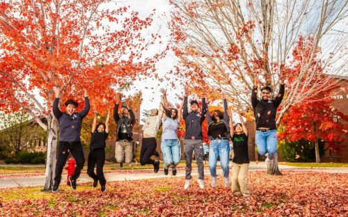 Students throw red, orange, and yellow autumn leaves in the air while jumping atop the grass in TMCC's plaza.