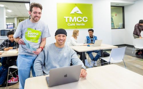 Two students smile while standing and seated at a table, holding a textbook and laptop in hand among others in front of the TMCC Cafe Verde.