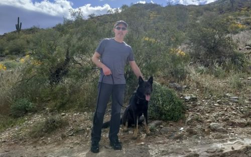 Damien Ennis and his dog, Baskerville, hike a trail in Nevada surrounded by a blue sky, sagebrush, green bushes, yellow goldenrod flowers, and cacti.