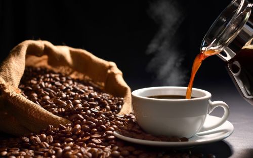 Coffee beans spill out of a cloth bag next to a steaming mug of coffee being poured.