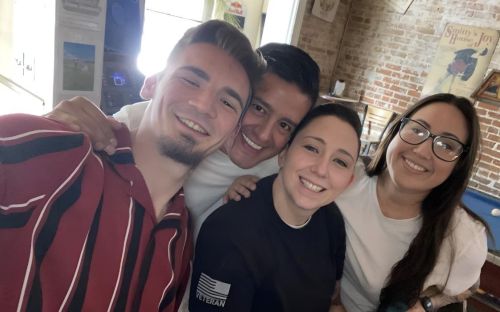 Sam McGuire and three friends smile for a group photo with a blue billiard table, a white cue ball resting atop, and brick walls in the background.