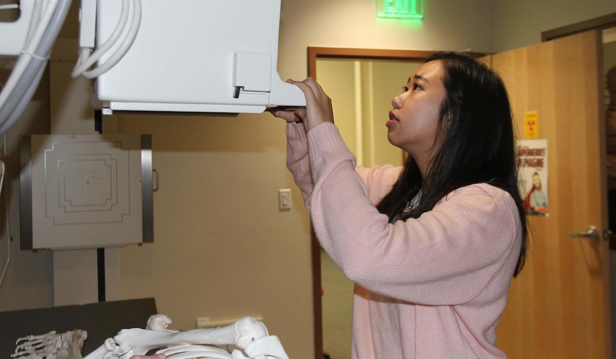 A Radiologic Technology student examines an X-ray machine before taking an image of the demonstration skeleton lying on the table.