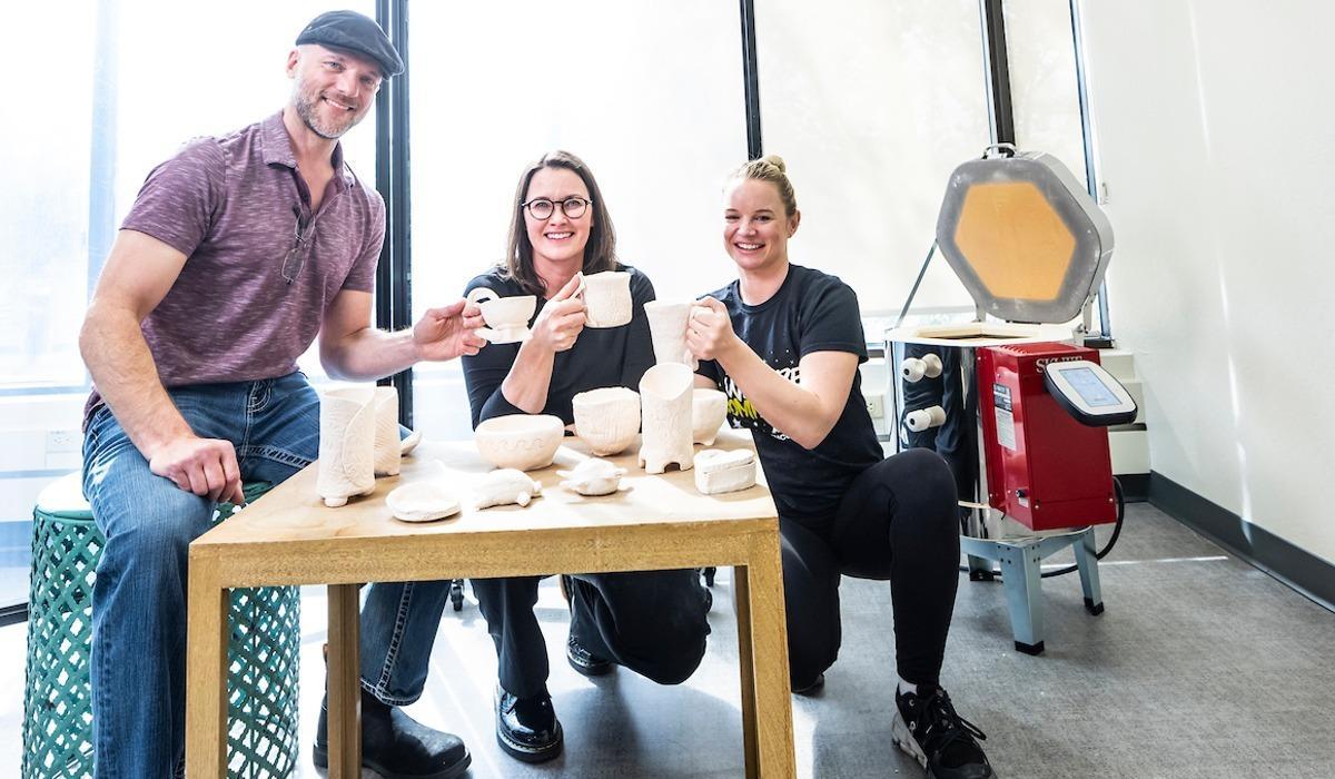 Three EPIC pottery class students sit smiling at a beige wooden table covered with ceramic objects while presenting their cup creations to the camera.