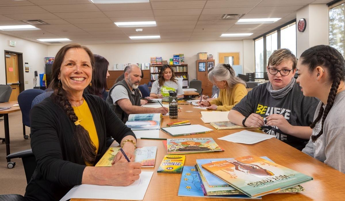 Dr. Micaela Rubalcava smiles at the camera, sitting at a lengthy beige desk with her students alongside children's books, colored pencils, and paper.