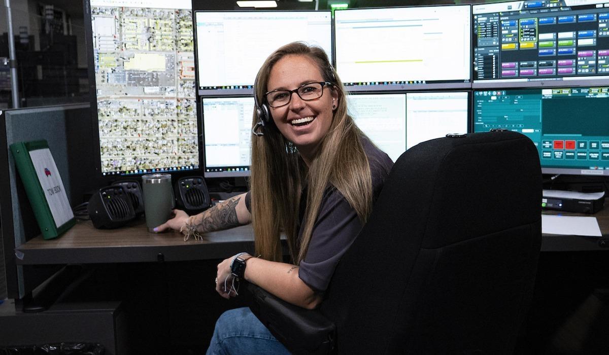 An emergency dispatch responder smiles while sitting behind her six computer screens, monitoring incoming or outgoing calls.