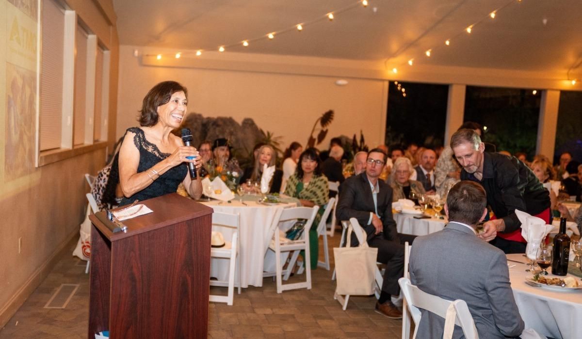 Dr. Cecilia Vigil gives her acceptance speech to a crowd sitting in white chairs and tables with strings of warmly lit bulbs above.