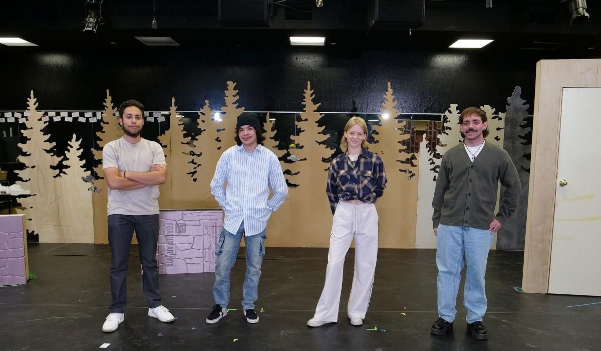 Four theatre students pose, smiling on a "Nearly, Washington" set piece with beige trees in the background, a door, and overhanging stage lights.