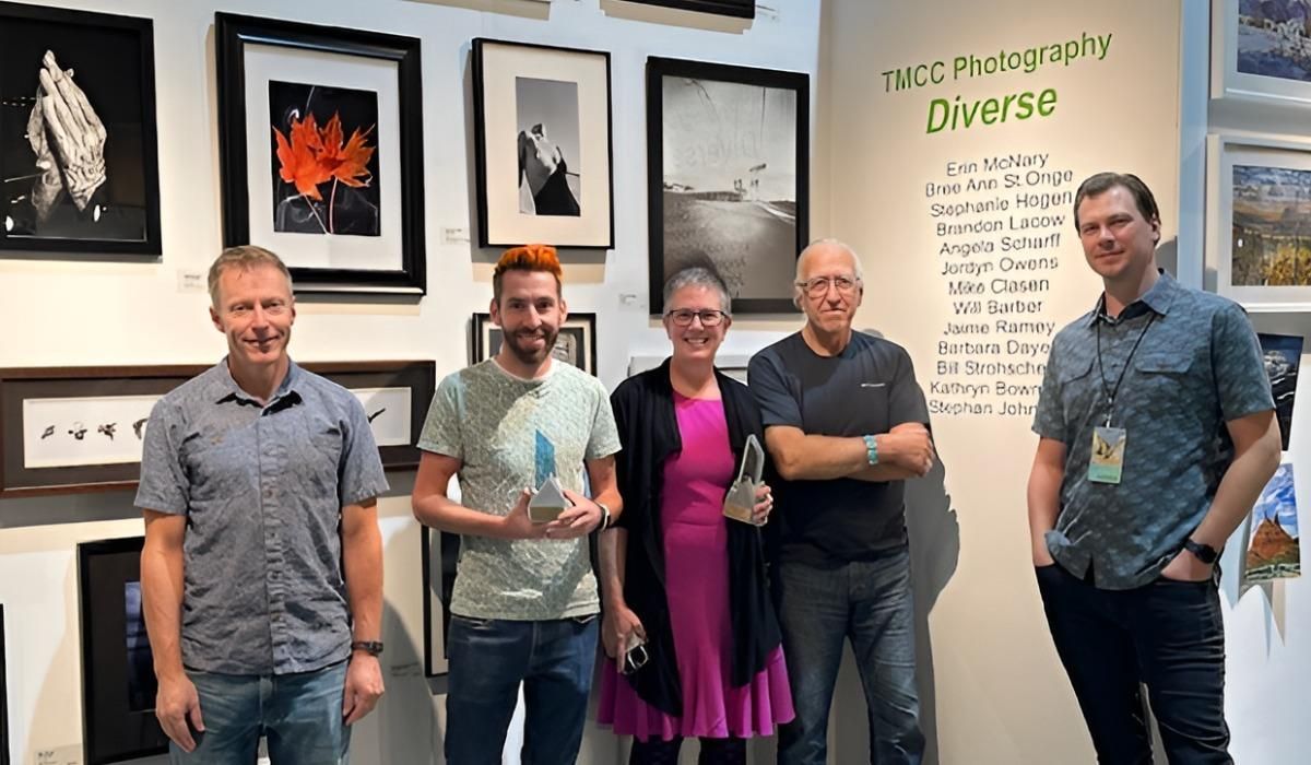 Candace Garlock stands beside colleagues in the Visual and Performing Arts Program, holding her award. Mesmerizing imagery is behind her on the wall.