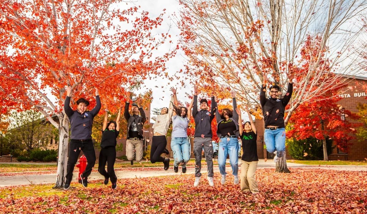 Students throw red, orange, and yellow autumn leaves in the air while jumping atop the grass in TMCC's plaza.