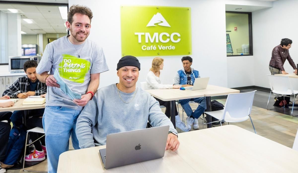 Two students smile while standing and seated at a table, holding a textbook and laptop in hand among others in front of the TMCC Cafe Verde.