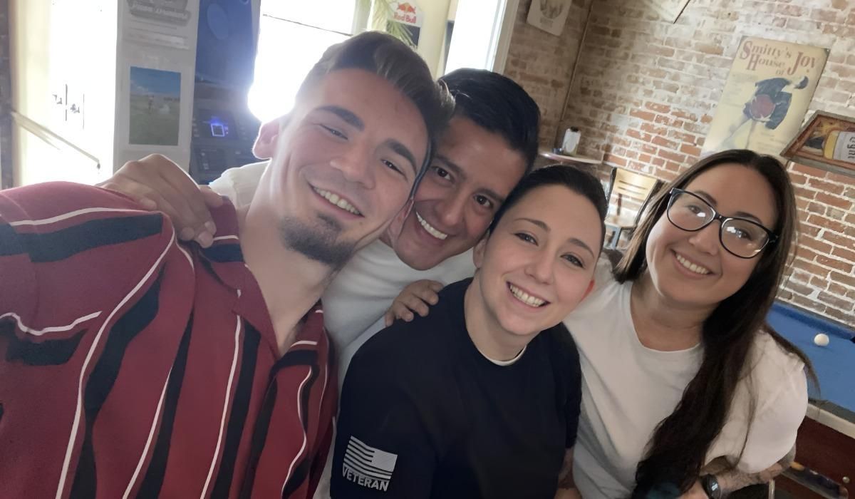 Sam McGuire and three friends smile for a group photo with a blue billiard table, a white cue ball resting atop, and brick walls in the background.