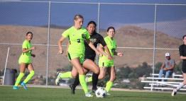 A Mighty Lizard Women's Soccer player, wearing a neon green jersey, competes against a female opponent decked in black as they fight for the ball on the pitch of TMCC's Soccer Field.