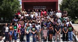 A large group of smiling Summer Bridge students stand outside the Red Mountain Building entrance by the plaza for a photo together.