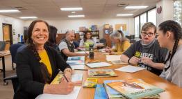 Dr. Micaela Rubalcava smiles at the camera, sitting at a lengthy beige desk with her students alongside children's books, colored pencils, and paper.