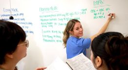 Three nursing students read and write instructions on a whiteboard for implementing medications at home, in the hospital, and in labs.