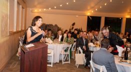 Dr. Cecilia Vigil gives her acceptance speech to a crowd sitting in white chairs and tables with strings of warmly lit bulbs above.