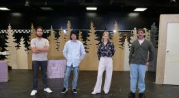 Four theatre students pose, smiling on a "Nearly, Washington" set piece with beige trees in the background, a door, and overhanging stage lights.