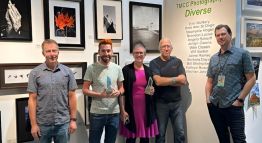 Candace Garlock stands beside colleagues in the Visual and Performing Arts Program, holding her award. Mesmerizing imagery is behind her on the wall.