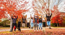 Students throw red, orange, and yellow autumn leaves in the air while jumping atop the grass in TMCC's plaza.