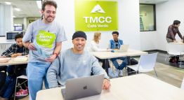 Two students smile while standing and seated at a table, holding a textbook and laptop in hand among others in front of the TMCC Cafe Verde.