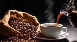 Coffee beans spill out of a cloth bag next to a steaming mug of coffee being poured.