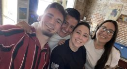 Sam McGuire and three friends smile for a group photo with a blue billiard table, a white cue ball resting atop, and brick walls in the background.